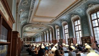 Innenansicht der prächtigen Bibliothek der Sorbonne mit lernenden Studierenden an Tischen. Decken und Wände mit Stuck und Ornamenten, hohe Sprossenfenster, auf den Tischen weiße Leselampen.