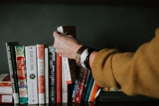 Eine Person, von der nur der Arm mit ockergelbem Pullover und Armreifen zu sehen ist, zieht ein Buch aus einem Bücherregal.
