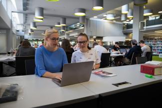 Zwei Studentinnen sitzen in der Universitätsbibliothek Heidelberg an einem Tisch mit aufgeklapptem Laptop, hinter ihnen andere lernende Studierende