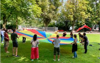 In einem Park halten ca. 20 im Kreis aufgestellte Menschen einen bunten Fallschirm zwischen sich gespannt, auf dem ein Gegenstand in Bewegung gesetzt wird. | © Yvo Wüest