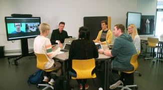 Ein Dozent sitzt mit 5 Studierenden an einem aus Dreieckstischen gebildeten Tisch; links hinten auf dem Smartscreen ein zugeschalteter Kollege.