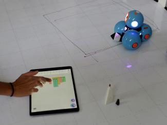 Auf weißem Fliesenboden liegt ein Tablet, mit dem eine Person, von der die Hand sichtbar ist, den blauen Roboter DASH! steuert.