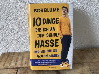 Auf einem hölzernen Tisch steht angelehnt an die Wand das Buch „10 Dinge, die ich an der Schule hasse und wie wir sie ändern können“ von Bob Blume. Die gebundene Ausgabe ist in Gelb gehalten und zeigt neben dem Titel ein Ganzkörperbild des Autors in gelbem Sweater und gelben Sneakers.