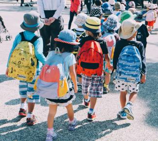 Eine von hinten abgelichtete Gruppe Grundschüler:innen mit Rucksäcken und Sonnenhüten auf dem Weg zur Schule