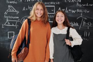 Zwei Schülerinnen mit Taschen über der Schulter stehen vor einer Tafel, auf der geometrische Formen abgebildet sind.