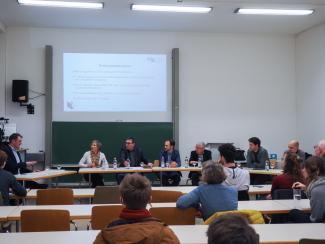 In einem Hörsaal der Neuen Universität sitzen die 6 Teilnehmenden der Podiumsdiskussion vor dem Publikum; links im Bild Dr. Florian Grafl.  