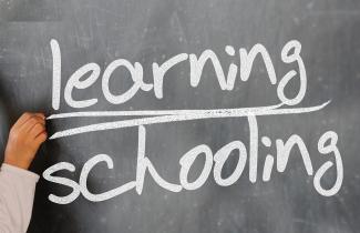 Auf einer schwarzen Tafel schreibt eine Hand die Worte „learning“ und „schooling“, wobei „learning“ doppelt unterstrichen ist.