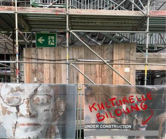 Das Bild zeigt ein eingerüstetes Gebäude, davor ein Bauzaun, der mit dem Plakat einer zerbrochenen Büste und dem Hinweis "UNDER CONSTRUCTION" versehen ist. Darüber in Rot: KULTURELLE BILDUNG: 