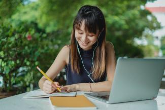 Eine junge Frau sitzt lächelnd draußen an einem Tisch. Sie schreibt in ihren Notizblock. Vor ihr ein aufgeklappter Laptop.