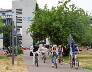 5 Studierende radeln durch den Campus im Neuenheimer Feld, hinter ihnen der Neubau der Pädagogischen Hochschule.