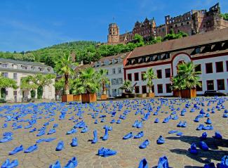 Auf dem Heidelberger Marktplatz stehen hunderte blau bemalte Schuhe, im Hintergrund das Heidelberger Schloss bei blauem Himmel. 