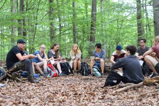 Studierende sitzen mit einem Dozenten im Kreis auf dem Waldboden, im Hintergrund Bäume.