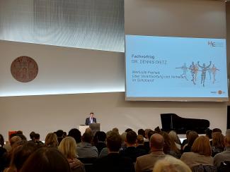 Dr. Dennis Dietz bei seinem Fachvortrag „Wertvolle Freiheit. Über Verantwortung und Vertrauen im Schulberuf“ am Rednerpult. Neben ihm die Leinwand mit Vortragstitel, vor ihm Publikum.