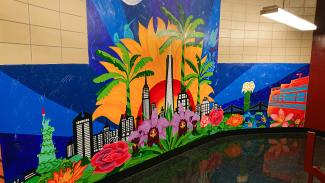 Das Bild zeigt ein in tropischen Farben gehaltenes, handgemaltes New York-Wandbild in einem gekachelten High-School-Flur.