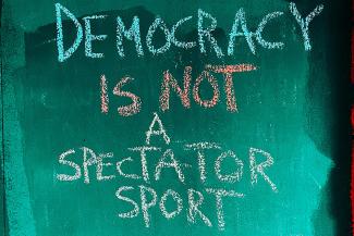 Auf einer petrolfarbenen Tafel steht mit bunter Kreide in Versalien "DEMOCRACY IS NOT A SPECTATOR SPORT".