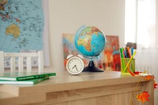 Das Bild zeigt einen hölzernen Schreibtisch mit Globus, Uhr, Stifteköcher und Büchern. Im Hintergrund ein Stuhl vor einer Weltkarte.