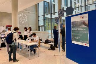 Im Foyer des Mathematikons stehen Tische für das Tagungsbüro des Heidelberger Informatiklehrkräftetags, an denen Mitglieder des HSE-Teams die Unterlagen für die neu ankommenden Teilnehmenden ausgebreitet haben. Eine Teilnehmerin nähert sich gerade den Tischen und dem Personal des Tagungsbüros. Rechts im Vordergrund sieht man eine Stellwand, auf der das Plakat des HILT hängt.
