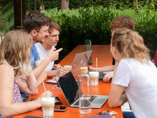 Eine gemischte Gruppe Studierender im Gespräch an einer Bierbank, vor sich Kaffee und Laptops.
