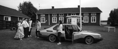 Filmstill in Schwarz-Weiß, das 5 Menschen vor einem alten Auto zeigt; links und im Hintergrund ein Haus.