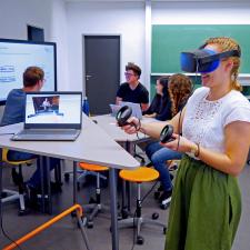 Innenansicht des neuen Lab-Standorts P18: Neben einem dreieckigen Stehpult nutzt eine Studentin das VR-Set; im Hintergrund sind vor einem Smartcreen weitere Personen um einen flexiblen Arbeitsplatz gruppiert.