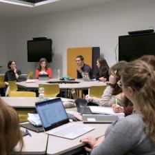 Eine Gruppe Studierender sitzt im Seminarraum P18 an kreisförmig angeordneten Tischen, auf denen Laptops und Arbeitsmaterialien liegen. Ein Student spricht gerade, die anderen hören zu.