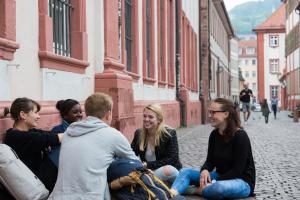 Eine gemischte Gruppe Studierender sitzt in der Heidelberger Altstadt vor einem Seminargebäude auf dem Boden