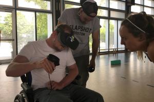 In einer Turnhalle treibt ein Student im Rollstuhl Sport mit VR-Brille und Controllern, 2 Studierende unterstützen