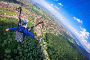 Junge Person (von hinten aufgenommen) schwebt im Tandem-Paragliding-Flug über Wald- und Stadtlandschaft, die Arme ausgebreitet