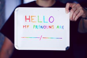 Eine junge Person, deren Gesicht nicht erkennbar ist, hält ein Pappschild hoch, auf dem in Regenbogenfarben steht: „HELLO my pronouns are -/-"