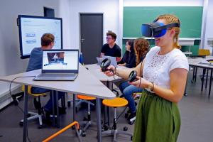 Innenansicht des neuen Lab-Standorts P18: Neben einem dreieckigen Stehpult nutzt eine Studentin das VR-Set; im Hintergrund sind vor einem Smartcreen weitere Personen um einen flexiblen Arbeitsplatz gruppiert.