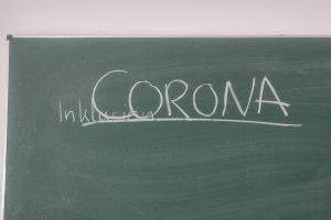 Auf einer grünen Tafel steht in großen Lettern unt unterstrichen das Wort „CORONA“; kleiner darunter und aufgrund der Nähe zu CORONA schlecht zu sehen das Wort „Inklusion“