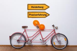 Ein orangefarbenes Tandem lehnt an einer Wand, am hinteren Lenker sind 2 orangefarbene Luftballons angebunden. Darüber der Schriftzug „Lehr:werkstatt“ in Orange, außerdem 2 Wegweiser: Auf dem einen steht „Heidelberg“, auf dem anderen „Praxiserfahrung“. 