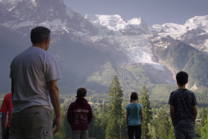 Szenenbild aus der Dokumentation Teachers for Life. Eine Lehrkraft und mehrere Schüler:innen sind von hinten zu sehen wie sie auf ein Bergmassiv mit schneebedeckten Gipfeln schauen