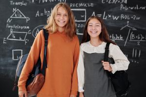 Zwei Schülerinnen mit Taschen über der Schulter stehen vor einer Tafel, auf der geometrische Formen abgebildet sind.