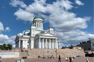 Man sieht den Dom von Helsinki bei schönem Wetter. Auf der großen Treppe und dem Platz davor spazieren einige Menschen.