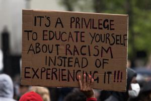 Schild auf einer Demonstration mit dem Text: "It's a privilege to educate yourself about racism instead of experiencing it!!!", hochgehalten von einer schwarzen Hand.