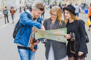 Ein junger Mann zeigt 2 jungen Touristinnen in einer Fußgängerzone etwas auf dem Stadtplan.
