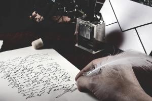 Eine Hand schreibt mit einem weißen Federkiel einen Brief; daneben Tintenfass und Umschlag.