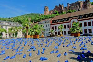 Auf dem Heidelberger Marktplatz stehen hunderte blau bemalte Schuhe, im Hintergrund das Heidelberger Schloss bei blauem Himmel. 
