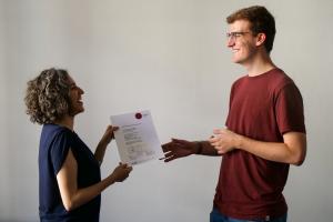 Ein Student nimmt von einer Mitarbeiterin des HSE-Prüfungsamts sein Zeugnis entgegen, beide lächeln.
