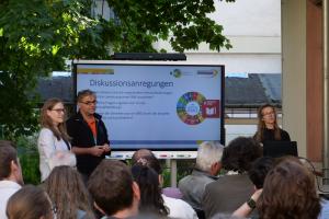 Teresa Ruckelshauß, Prof. Dr. Alexander Siegmund und Dr. Johanna Weselek vor einem Smartboard auf der Terrasse der HSE. Im Hintergrund ein Baum, im Vordergrund die Hinterköpfe des Publikums. Auf den Screen die Überschrift "Diskussionsanregungen" mit 3 Leitfragen.