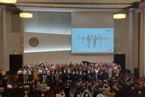 Die Absolvent:innen der Studiengänge Master of Education Profillinie Lehramt Gymnasium sowie Master of Education Gerontologie, Gesundheit und Care auf der Bühne der Neuen Aula der Universität Heidelberg. In den Händen halten sie eine HSE-Mappe. Auf die Leinwand hinter ihnen ist eine stilisierte Skizze projiziert, die Läufer:innen beim Zieleinlauf zeigt. Im Vordergrund sind Angehörige zu erkennen, die mit ihren Smartphones Bilder machen.