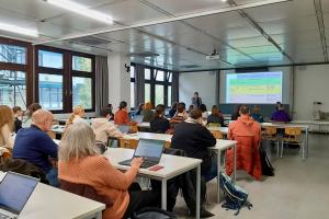 Teilnehmende des Workshops "KI-Unterstützung anhand aktueller Tools für Forschung und Unterricht" sitzen in einem Seminarraum im Neubau der Pädagogischen Hochschule Heidelberg und schauen nach vorne auf ein Projektionsfläche. Neben der Projektionsfläche steht einer der Referenten der Veranstaltung, der HSE-Mitarbeiter Dr. Thomas Renkert.