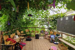 Auf dem Dielenboden einer dschungelartig begrünten Veranda sind Holzstühle auf eine Tafel ausgerichtet, auf der Lebensmittelnamen in Spalten sortiert sind. Auf dem Boden ein Korb mit CO2-Schild sowie weitere Materialien..  