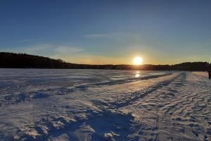 Das Foto zeigt einen klaren, blauen Himmel und eine weite Schneefläche, die durch die untergehende Sonne in ein kühles, aber gleißendes Licht getaucht wird. In der verschneiten Fläche zeichnen sich schemenhaft eine Eisfläche und Skispuren ab. Menschen spazieren über dem zugefrorenen See in Jyväskylä, der im Winter als Sport- und Freizeitort genutzt wird.