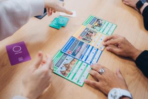 Auf einem Holztisch sind 4 Spielkarten zu sehen, die sich mit den Nachhaltigkeitszielen befassen. 5 Hände interagieren mit den Karten.