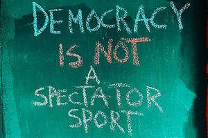 Auf einer petrolfarbenen Tafel steht mit bunter Kreide in Versalien "DEMOCRACY IS NOT A SPECTATOR SPORT".