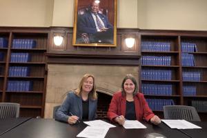 KerryAnn O'Meara, Provost und Dean des Teachers College, und Prof. Dr. Frauke Melchior, Rektorin der Universität Heidelberg, unterzeichnen an einem Schreibtisch im Teachers College das Memorandum of Understanding. Im Hintergrund Regale mit blauen Büchern, darüber ein Porträt.