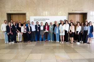 Gruppenbild der Vertreterinnen der lehrkräftebildenden Hochschulen Baden-Württembergs vor dem Senatssaal der Universität Stuttgart. Im Hintergrund der Schriftzug "Verbund Lehrkräftebildung Baden-Württemberg" auf weißem Grund.