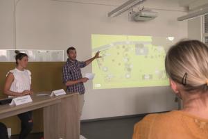 Ein Student und eine Studentin bei einer Präsentation. Der Student deutet auf eine projizierte Karte. Im Vordergrund eine Zuhörerin.